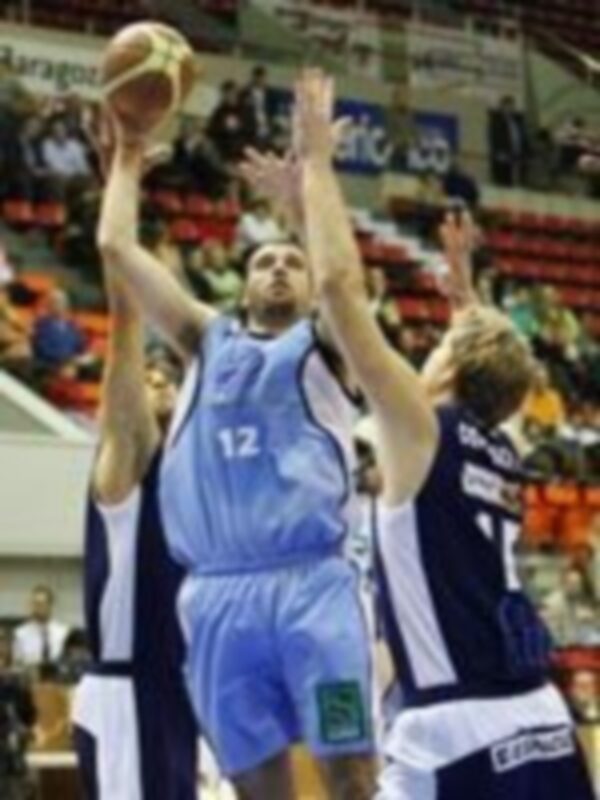 Un jugador de baloncesto en acción, con el número 12 en su camiseta, lanzando una canasta mientras otro jugador lo observa. El fondo muestra un estadio lleno de espectadores y la portería del equipo contrario.