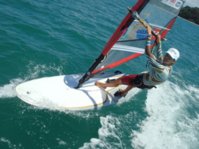 Un windsurfer disfruta de su deporte en el agua azul, con una vela roja y blanca en la parte superior. El windsurfer está usando un traje de surf multicolor y una gorra blanca, mientras se desplaza sobre la superficie del agua.