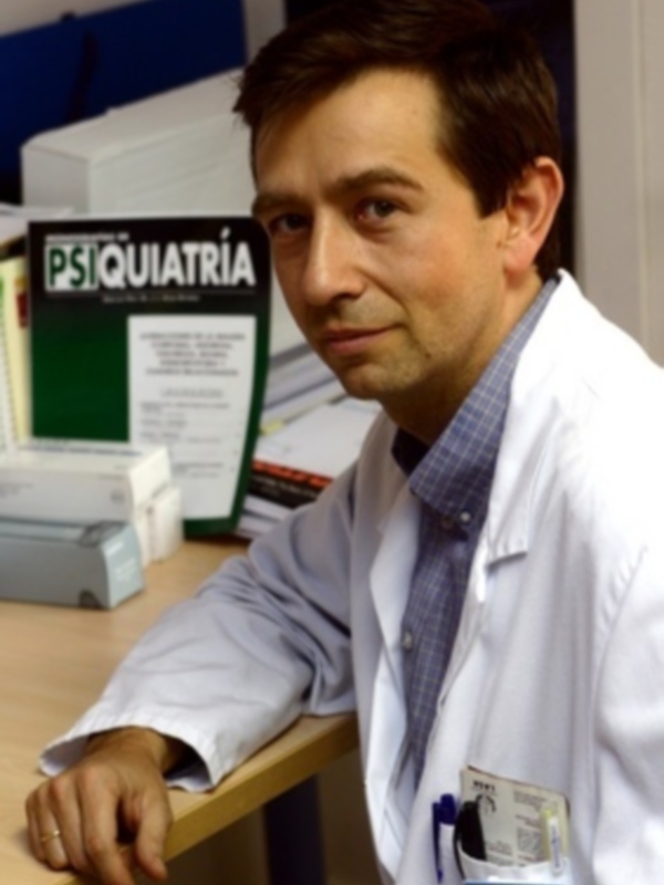 Médico en uniforme blanco, sentado en una oficina médica con libros y documentos al fondo.