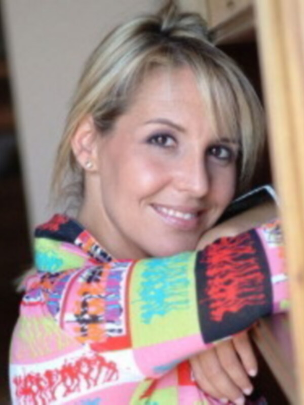 Una mujer con cabello largo y rubio, vistiendo una camiseta de colores brillantes. La imagen parece estar tomada en un entorno urbano, posiblemente una calle o área de trabajo.