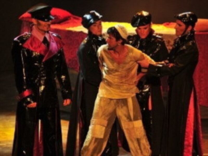 Una escena teatral con un hombre en el centro, rodeado por cuatro personas vestidas de negro y rojo. La escena parece ser parte de una representación teatral o musical, con un fondo rojo y escenario iluminado.