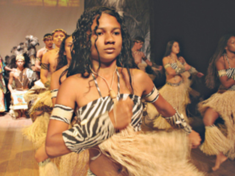 Una joven en el centro, vestida con un atuendo tradicional de rayas negras y blancas, posa mientras otros bailarines con vestimentas similares se encuentran detrás. El ambiente es festivo y lleno de color, con una pista de baile en el fondo.
