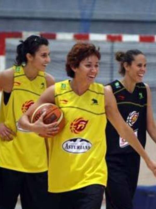 Las jugadoras de baloncesto son sonrientes y están en una cancha deportiva. La imagen muestra a tres jugadoras, dos de ellas con uniformes amarillos y una en negro. La jugadora central sostiene un balón de baloncesto.
