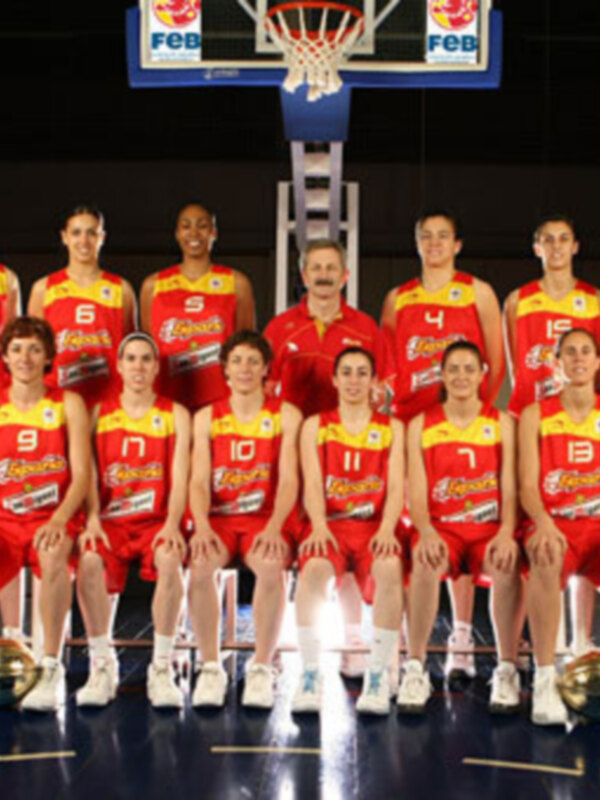 Equipo de baloncesto posando en cancha con camisetas rojas y amarillas, frente a una portería de baloncesto.