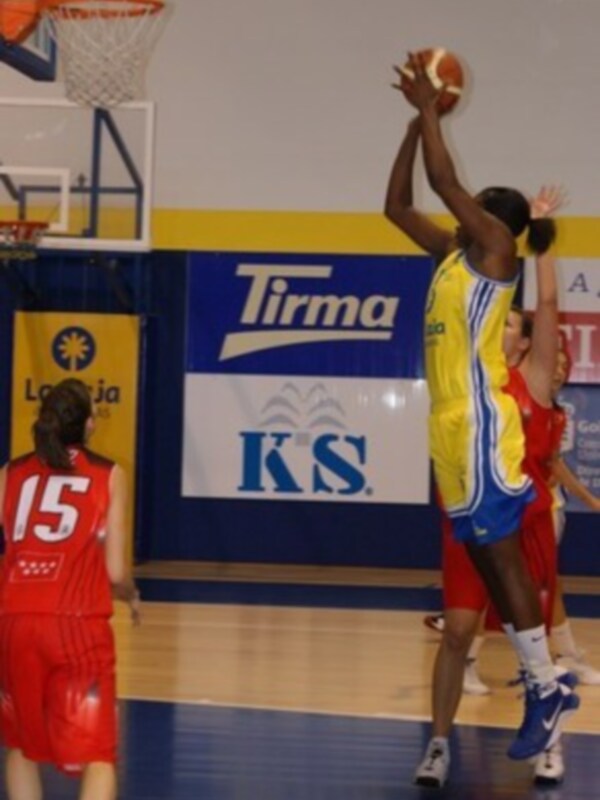 Un jugador de baloncesto en un uniforme amarillo y azul está a punto de lanzar el balón hacia la canasta, mientras que otros jugadores con uniformes rojos observan. El fondo muestra un anuncio de Tirma y KS en la pared del estadio.