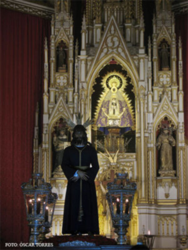 En una iglesia de estilo gótico, un hombre en traje negro se encuentra frente a una imagen de la Virgen María. La arquitectura dorada y el altar con estatuas religiosas completan la escena.