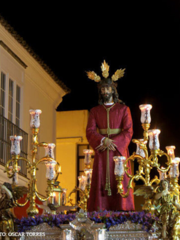 Escena nocturna con una estatua de Jesús en rojo, rodeada por candelabros dorados y flores. Fondo de edificio blanco con ventana.