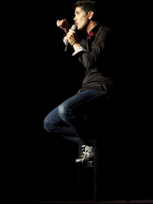 Un hombre joven con una camisa negra y un cuello rojo está cantando en un escenario oscuro. Se sienta con una pierna sobre la otra y sostiene un micrófono cerca de su boca.