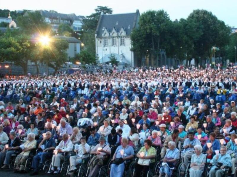 Una multitud asiste a un evento en un parque, con una iglesia al fondo y árboles circundantes. La gente está sentada en sillas de ruedas y otras posiciones, disfrutando del evento.
