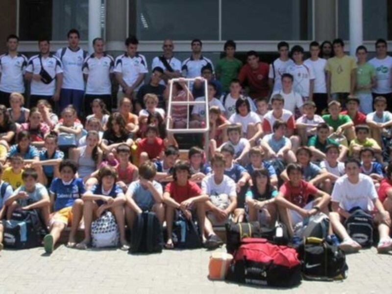 Grupo de personas sentadas en el suelo con maletas y mochilas. Fondo: edificio, palmera, ventana.