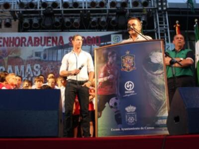 Un hombre hablando en un micrófono frente a una pantalla de la selección española, con una bandera y un escudo en el fondo.