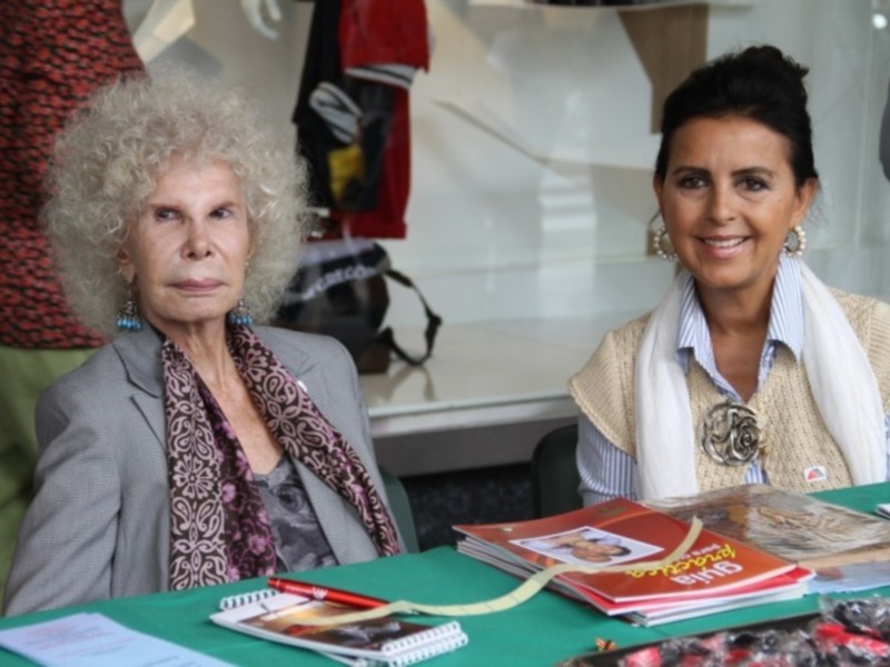 Dos mujeres sentadas en una mesa con libros y revistas. Una tiene cabello blanco, la otra tiene el cabello negro y lleva un pañuelo. En el fondo hay una mesa con bolsas y otros objetos.