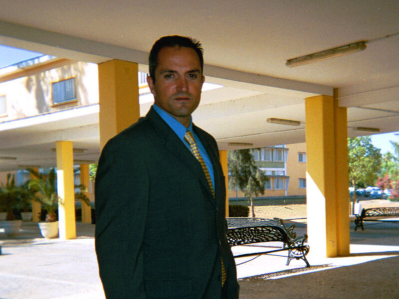 Un hombre viste un traje oscuro y una camisa azul con corbata amarilla, posando frente a un edificio de varios pisos con toldos en el exterior. La escena se desarrolla en un entorno urbano, con columnas amarillas y una mesa de metal en el fondo.