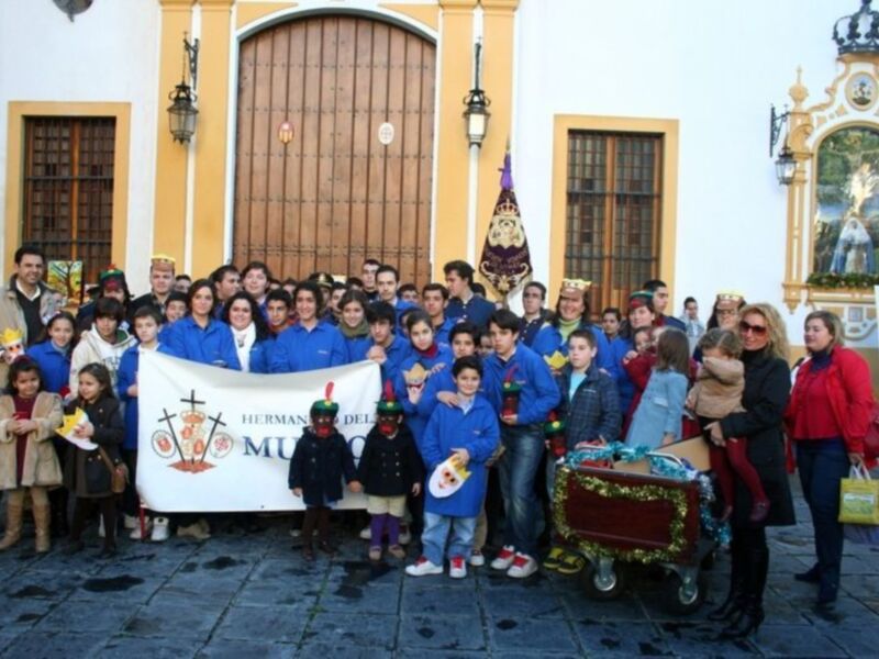 Grupo de personas con uniforme azul y bandera de 'Hermanos del Museo', posando frente a una fachada histórica decorada con motivos navideños.