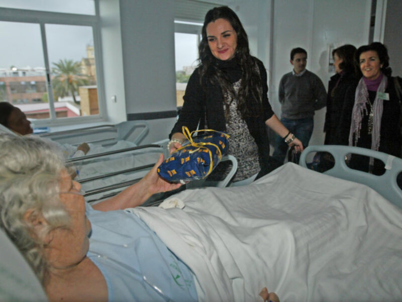 Una anciana en una cama de hospital, con una mujer joven a su lado sosteniendo un objeto amarillo y azul. En el fondo, se pueden ver ventanas con vistas a un edificio y árboles.