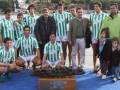 Equipo de fútbol en uniforme verde y blanco posando con una estatuilla de un balón, junto a varios espectadores y niños pequeños.