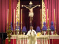 En el altar, un Cristo crucificado se destaca en la parte superior. A su alrededor, varios velas y flores azules adornan el espacio sagrado. Un sacerdote, vestido con un hábito blanco, se encuentra frente al altar. El fondo es una cortina roja que añade profundidad a la escena religiosa.