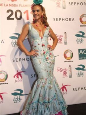 Una mujer con un vestido de flamenca azul claro y flores rojas, posando frente a una pared con logos de empresas.