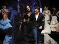 Una modelo con vestido negro y un hombre caminan por la pasarela de una feria de moda. En el fondo, se pueden ver imágenes de otros modelos en diferentes vestidos.