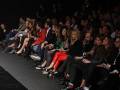Asistencia a una presentación de moda en un escenario con asistentes vestidos de manera elegante, algunas con zapatos de tacón alto y otros con vestidos formales.