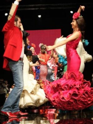 Una pareja baila flamenco en un escenario iluminado, con el hombre levantando la mano y la mujer con un vestido rojo volador.