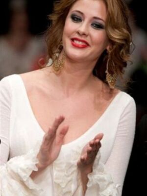 Una mujer con un maquillaje oscuro y labios rojos, lleva una vestimenta blanca con detalles en la manga. El fondo parece ser un escenario de alfombra roja, posiblemente una gala o evento formal.