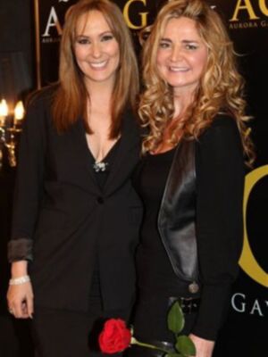 Dos mujeres sonrientes posan juntas, una con un vestido negro y la otra con un abrigo de cuero. La imagen está en un evento, con luces y una pancarta al fondo.
