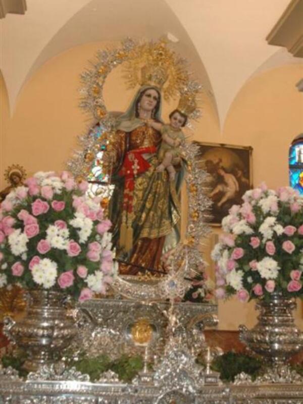 Imagen religiosa de una Virgen con un niño en sus brazos, rodeada por flores y adornos.