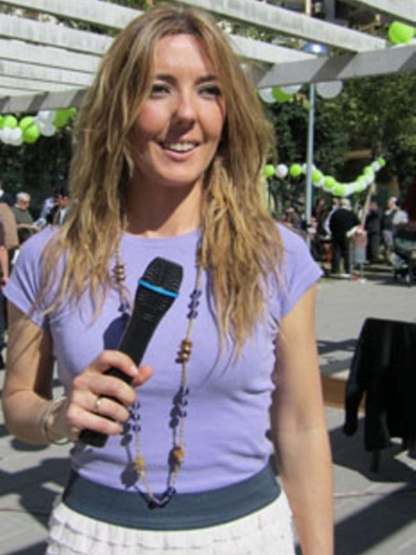 Una mujer con cabello largo y rizado, vestida con una camiseta púrpura y falda blanca, sostiene un micrófono. Fondo con decoración navideña y personas en el fondo.