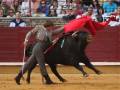 Un torero en chaqueta beige y pantalones grises, con un capote rojo, lucha contra un toro negro en una plaza de toros. La arena es de tierra y el público observa desde los muros rojos.