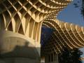 Una estructura arquitectónica moderna con un diseño de malla en forma de cúpula, típica del Metropol Parasol en Sevilla. La estructura es de color beige y tiene una gran altura, con un cielo azul claro en el fondo. En la parte inferior de la imagen se puede ver una escalera y un árbol, mientras que en el centro hay una bandera con los colores de la Unión Europea.