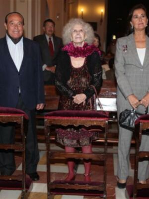Una mujer mayor con un vestido de color marrón y botas rojas, una mujer joven con un traje gris claro y una mujer mayor con un vestido negro y chaqueta de plumas, todos en una sala de audiencias.