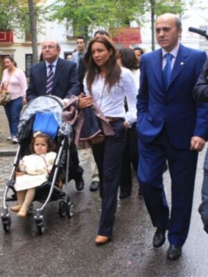 Una familia camina por una calle, con un hombre en traje azul y una mujer con vestido blanco. La niña está sentada en un carrito de bebé, y otros adultos están a su lado.