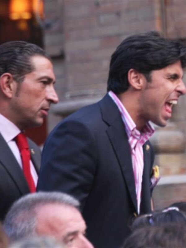 Dos hombres en trajes de negocios, uno con corbata roja y el otro con corbata rosa, posan juntos frente a un edificio con una chimenea en el fondo.