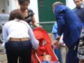 Una familia está en un parque, con una niña pequeña en un carrito de bebé. La madre está ayudando a la niña, mientras el padre observa. Hay otros adultos en la imagen, incluyendo un hombre en traje azul y una mujer con cabello largo. La escena parece ser en un día soleado, con edificios blancos y verdes al fondo.