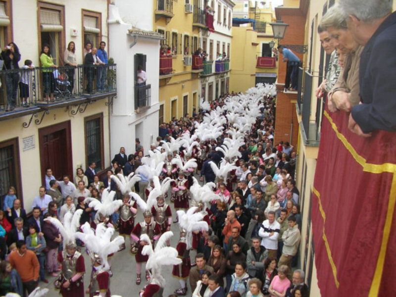 Una multitud en una calle de pueblo, con personas vestidas de blanco y plumas, seguidas por un grupo de adultos en balcones. La imagen muestra una celebración festiva con disfraces y decoraciones típicas de la cultura local.