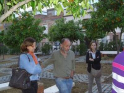 Una persona con una mochila habla a dos personas en un parque, rodeado de árboles y casas.