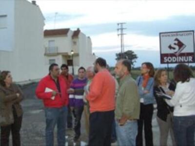 Una persona con un saco de compras se encuentra en el centro de una calle. A su alrededor, hay varias personas que parecen estar conversando o esperando algo. En el fondo se puede ver una señal de tráfico y un edificio blanco con ventanas. La escena parece ser en una zona urbana, posiblemente un barrio residencial.