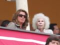 Dos mujeres y un niño observan desde una tribuna con una bandera roja. La imagen muestra una escena de espectadores en un evento deportivo o social.