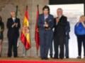 Una persona en traje azul sostiene un trofeo mientras otros individuos observan una ceremonia formal. Banderas españolas y un escudo nacional se destacan en el fondo, junto con una pantalla que muestra información sobre la celebración.