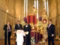 Una ceremonia religiosa en una iglesia con una estatua de Cristo en el centro, rodeada de velas y flores. Los participantes están vestidos formalmente, incluyendo un hombre en una chaqueta negra y corbata rosa. La atmósfera es solemnemente reverente.