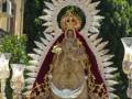 Imagen de la Virgen María con un manto rojo y dorado, rodeada de flores blancas y una corona dorada. La imagen está en un entorno con árboles verdes y una estructura de madera en el fondo.
