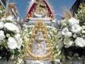 Una estatua de la Virgen María, rodeada de flores blancas y adornos dorados, en un entorno urbano con edificios al fondo.