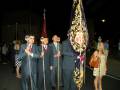 Personajes en trajes formales con palos y estandartes en una procesión nocturna.