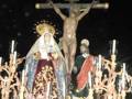 Una procesión religiosa nocturna con una estatua de Jesús crucificado en el centro, rodeado por figuras vestidas con trajes tradicionales y luces de velas que iluminan la escena. La estatua central es una representación de la Crucifixión, flanqueada por figuras que podrían ser personajes bíblicos o santos. La procesión está decorada con flores y velas, creando un ambiente de solemnidad y devoción.