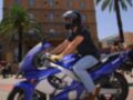 Una persona monta una motocicleta Yamaha azul en un entorno urbano con edificios y palmeras al fondo.