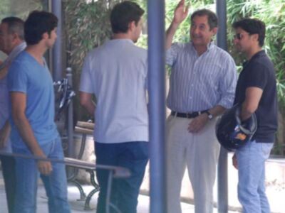 Un hombre en una camisa de rayas celebra con los brazos levantados mientras otros hombres lo saludan. Todos están en una zona de parque o jardín, con árboles y plantas al fondo.