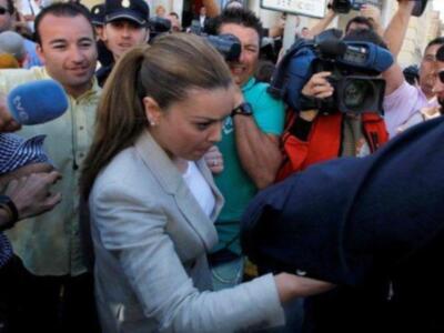 Una mujer en un abrigo blanco es rodeada por personas con cámaras y micrófonos. La escena parece ser en un lugar público, posiblemente una calle o plaza con edificios al fondo.