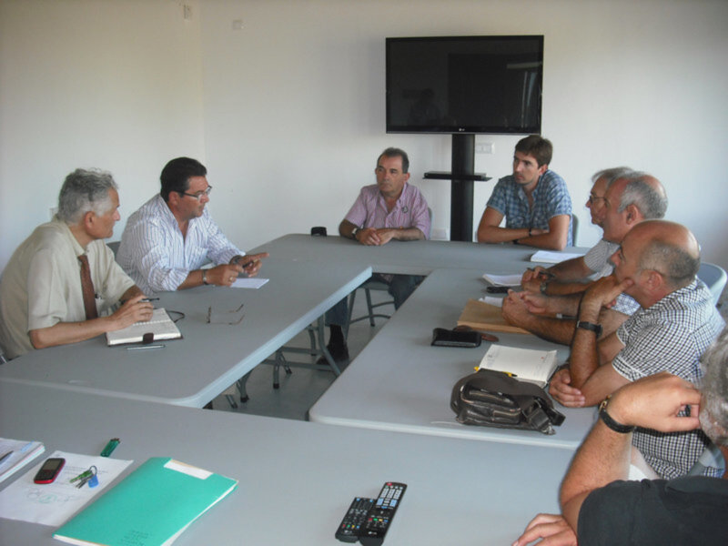 Gente reunida en una sala de conferencias, sentada alrededor de una mesa con documentos y dispositivos electrónicos.