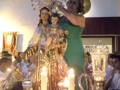 Una mujer sostiene una estatua de la Virgen María con un niño en sus brazos, rodeada por velas y una imagen de la Virgen María en el fondo.